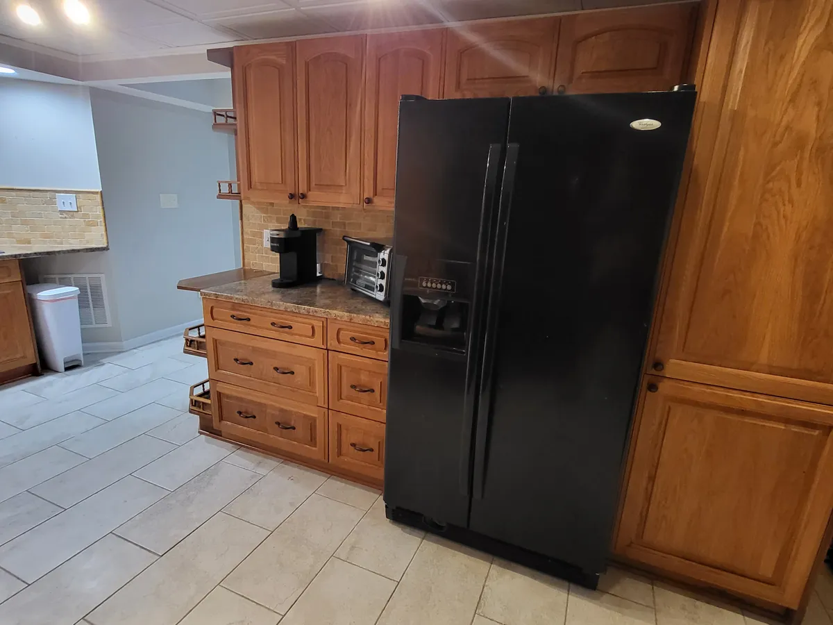 A kitchen setup featuring a sleek black refrigerator, wooden cabinets, granite countertop, and a coffee maker. The scene is practical and inviting, with plenty of space for meal preparation and storage, and hints at included amenities for daily living.
