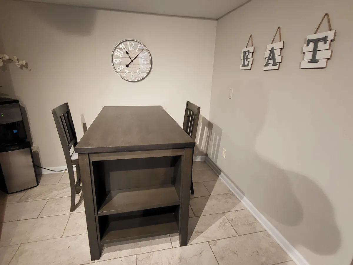 This image features a cozy dining area with a wooden table, wall clock, and lettered wall decor. Sunlight gently streams in, suggesting proximity to windows or perhaps a patio door, and the setting is styled for relaxed, everyday living. The mood is warm and inviting, hinting at moments spent enjoying morning coffee or casual meals.