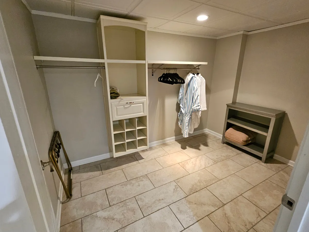 A well-organized walk-in closet with clothes neatly hung on hangers and a shelving unit for additional storage. The textured floor tiles and spacious layout highlight the ease of organizing personal belongings and the comfort of having ample storage space.