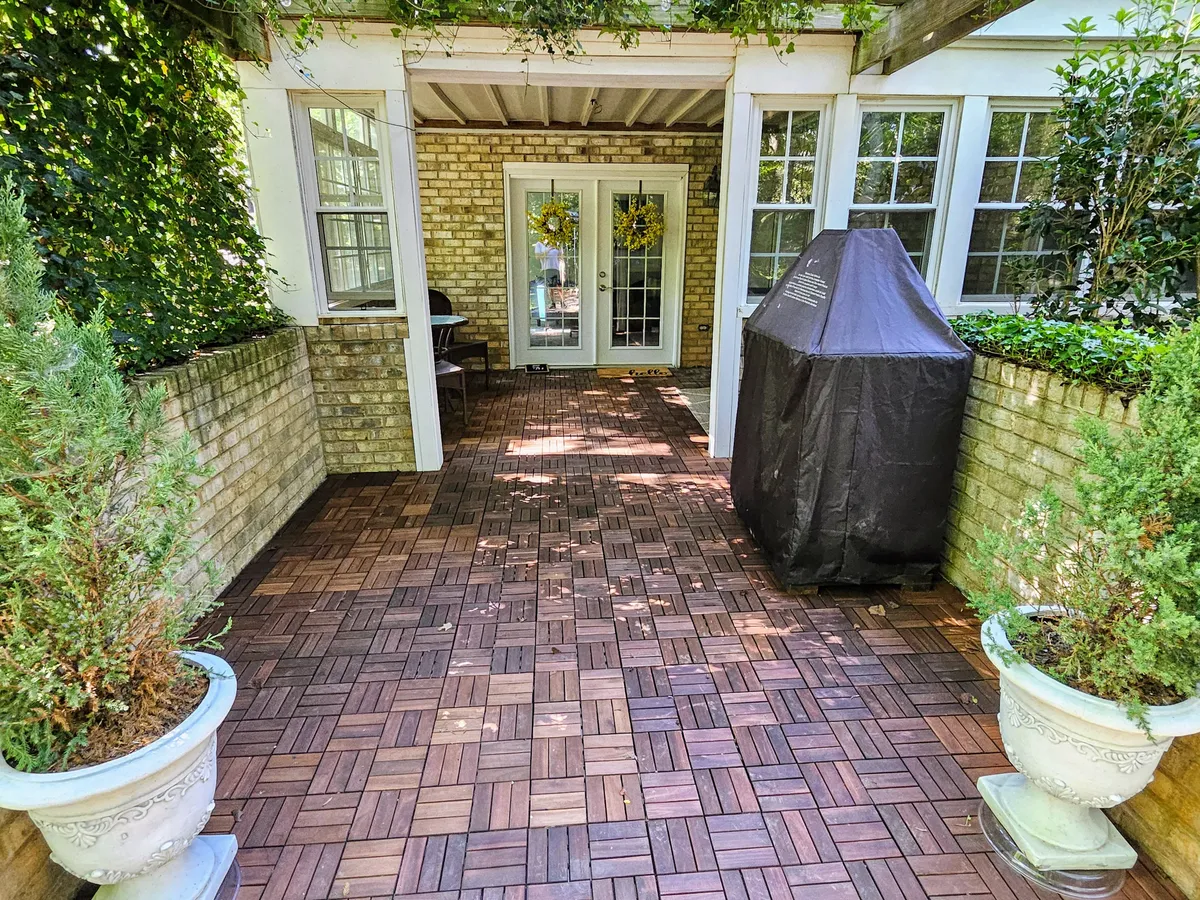 The image showcases the private garden-level patio of the apartment, featuring a sturdy brick wall, a glass door leading inside, lush potted plants, and a barbecue grill cover. The area is shaded and inviting, suggesting a tranquil spot for outdoor relaxation, morning coffee, or evening barbecues.
