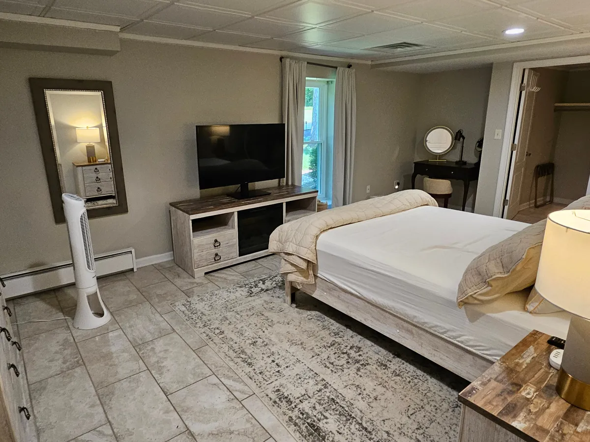 A tranquil bedroom featuring a comfortable bed with soft linens, a television for in-room entertainment, a dressing table for personal routines, and a large window letting in natural light. The space radiates comfort and relaxation, offering a restful atmosphere ideal for unwinding after a long day.