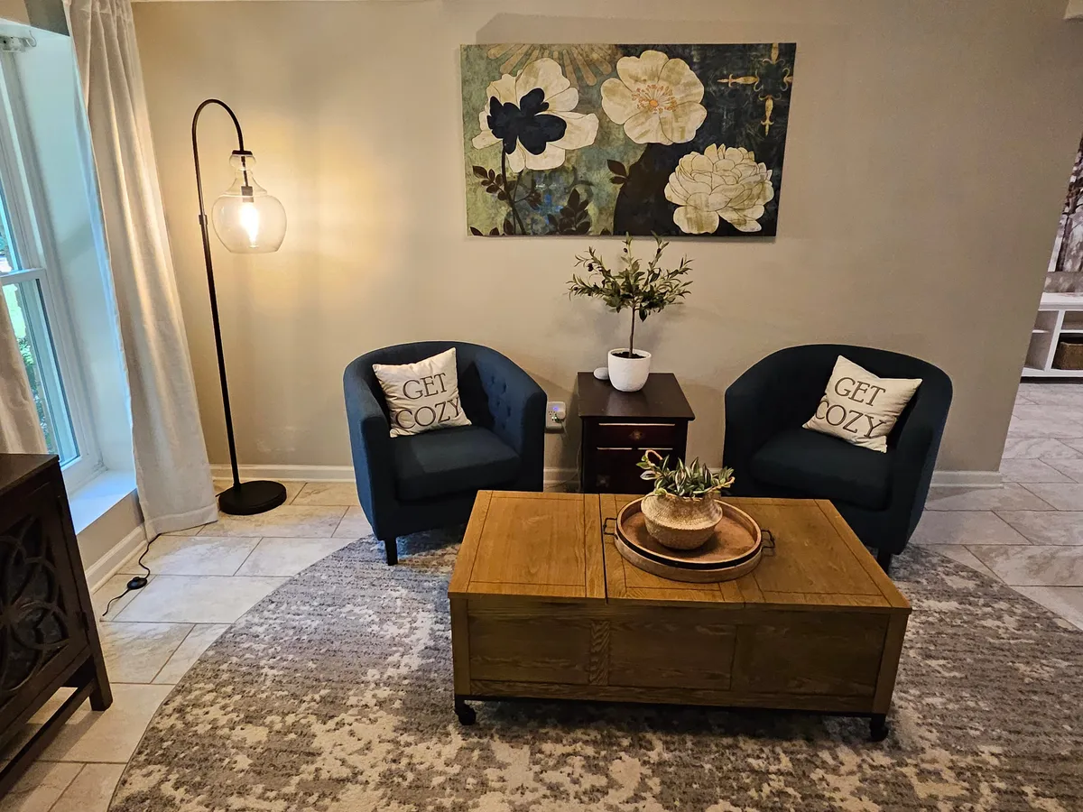 A pair of blue armchairs and a wooden coffee table set in a bright, cozy corner, illuminated by morning sun. The arrangement fosters intimate conversations or quiet reflection, and the visible sunlight hints again at a close connection to the outdoor patio or garden spaces.