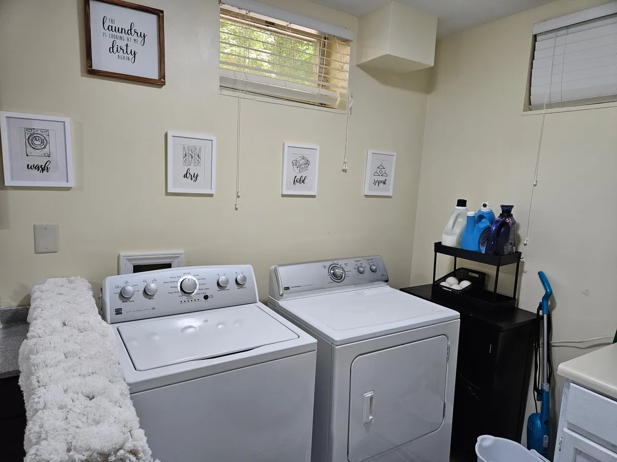 The image displays a well-kept laundry area featuring a modern washing machine and dryer, neatly arranged cleaning supplies, and framed wall art that adds a homely touch. The space is tidy, practical, and radiates a sense of comfort and utility.