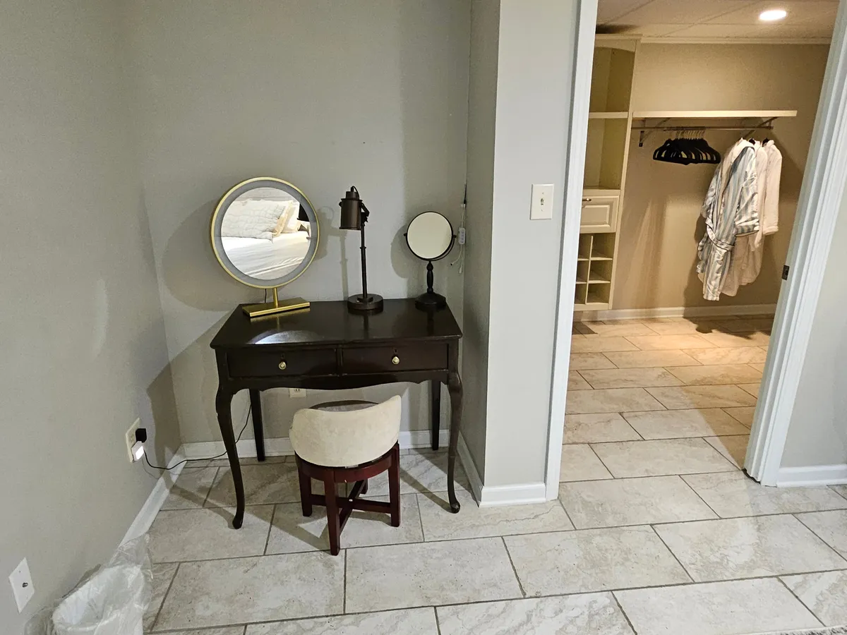 An intimate vanity corner complete with a round mirror, table lamp, and nearby closet creates a private haven for self-care. A comfortable stool and neatly arranged decor contribute to a sense of personal retreat and readiness for the day ahead.