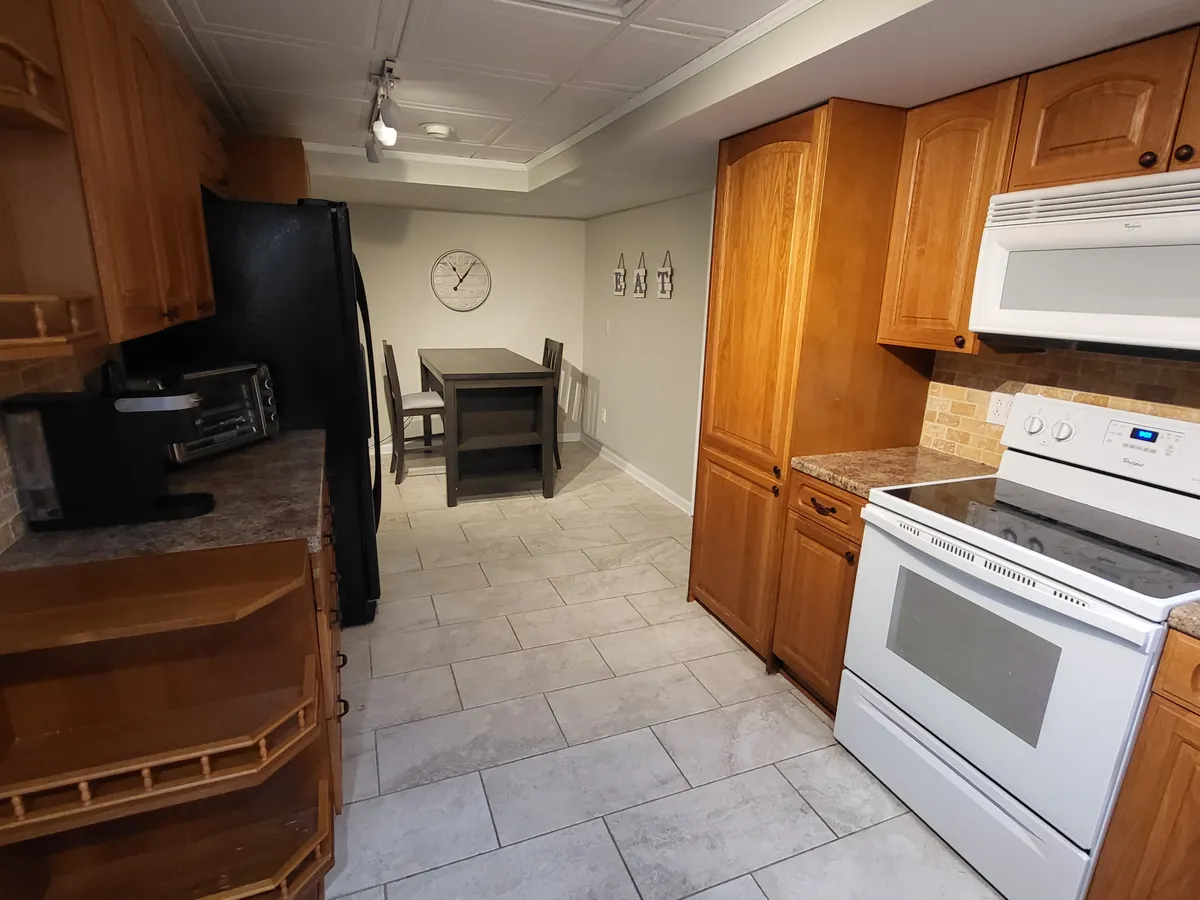 A cozy dining area features a wooden table set next to a classic kitchen with wooden cabinetry and white appliances. Decorative letters and a wall clock add a touch of personality, setting the scene for shared meals and everyday living. Natural light enhances the inviting, functional atmosphere, suggesting a seamless daily routine and meaningful connections at home.